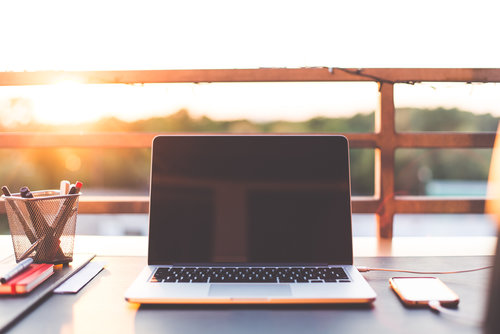 A laptop with a blank screen sits on an outdoor desk at sunset, flanked by a phone and a pen holder with pens and pencils. A railing and blurred landscape are visible in the background.