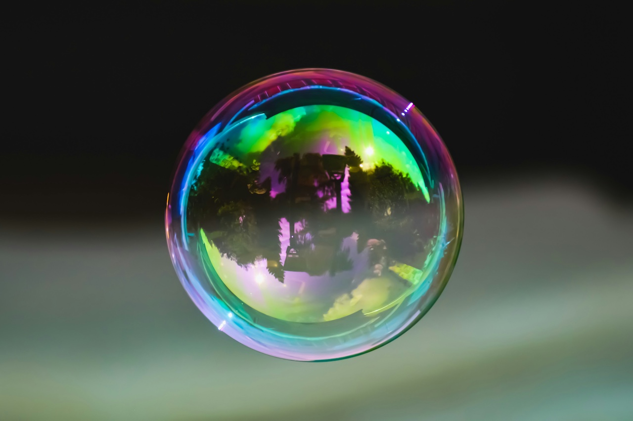 A close-up of a floating soap bubble with vivid rainbow colors and a distorted reflection of trees and buildings on its surface.