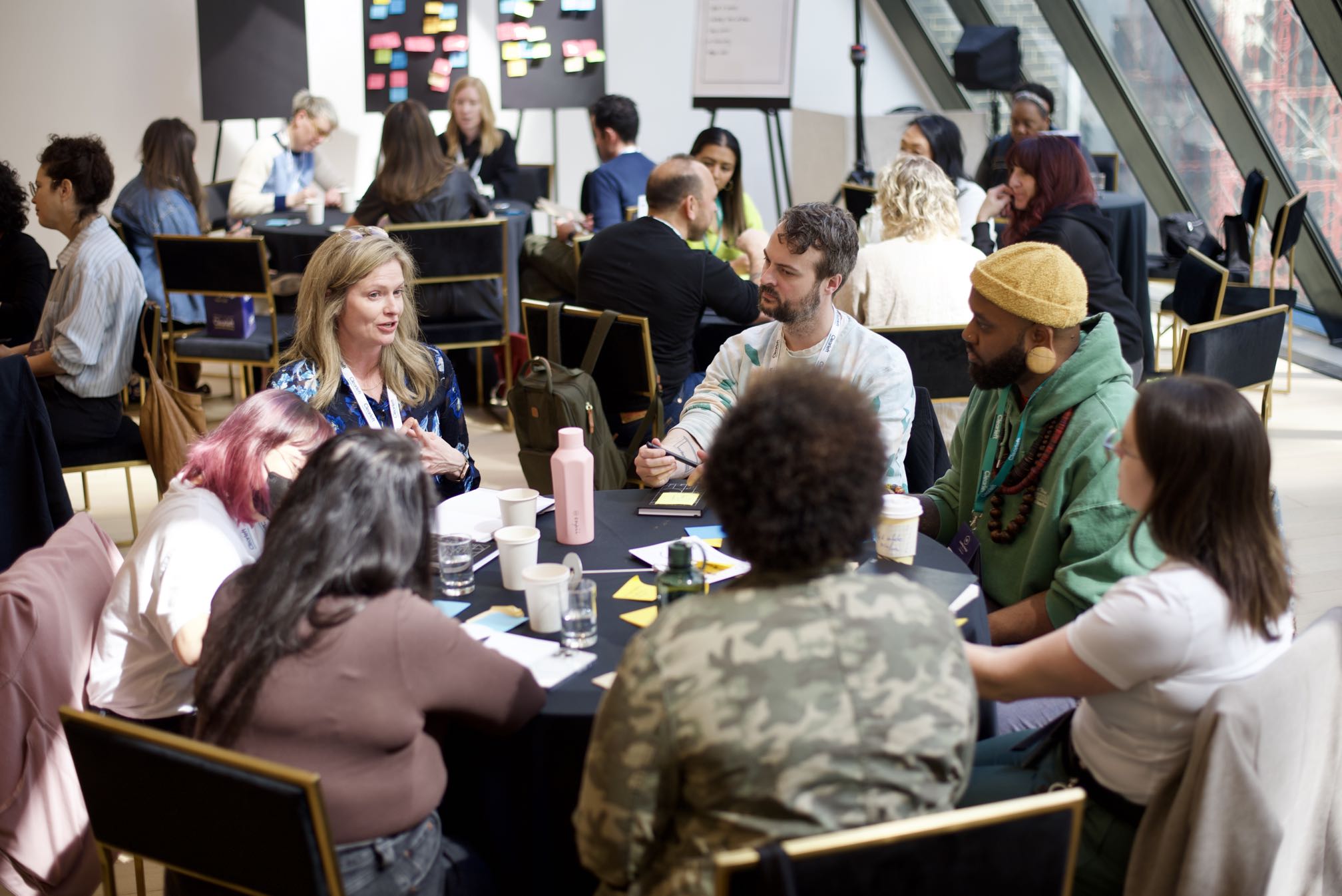 A group of people seated around round tables in a bright, modern conference space, engaged in discussion. Colorful sticky notes are visible on boards in the background, suggesting a collaborative or workshop setting.