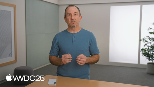 A man stands at a desk in a modern, minimal office setting, speaking or presenting. He wears a blue shirt and gestures with his hands. The Apple logo and “WWDC25” text in the lower-left corner indicate this is from Apple's Worldwide Developers Conference 2025. A smartphone lies on the desk in front of him.