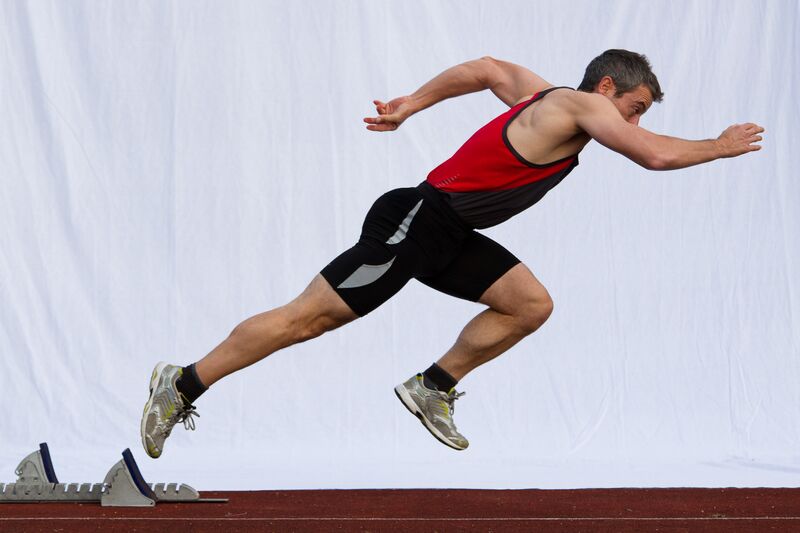 Male sprinter in a red and black athletic outfit launching from starting blocks on a track with a white background.