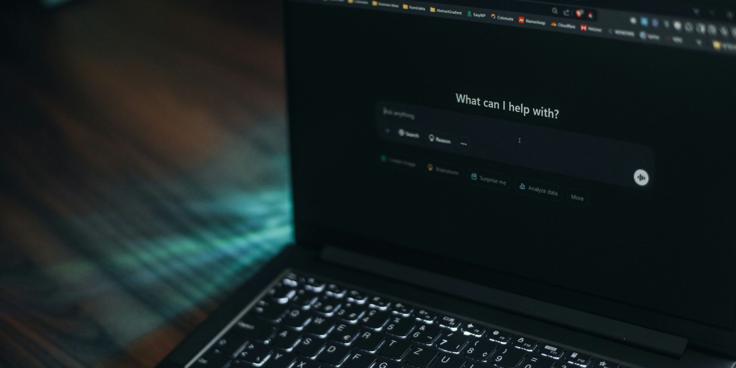 Close-up of a laptop keyboard and screen displaying a dark-themed AI assistant interface with the prompt 'What can I help with?' and a search input field.