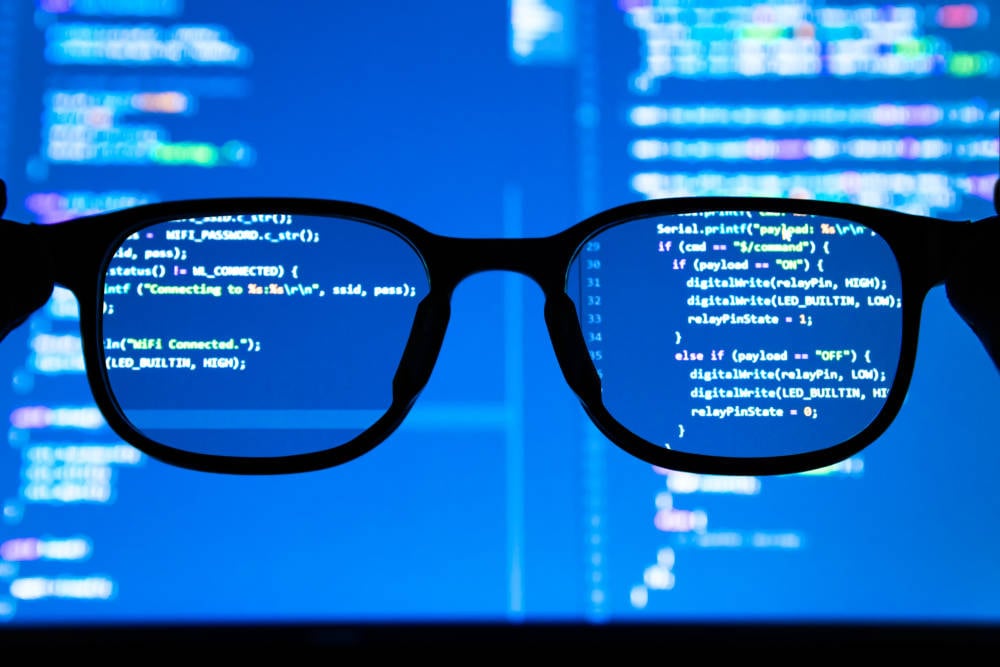 Black-rimmed eyeglasses in front of a computer screen, with sharp focus on code reflected in the lenses and blurred code in the background.