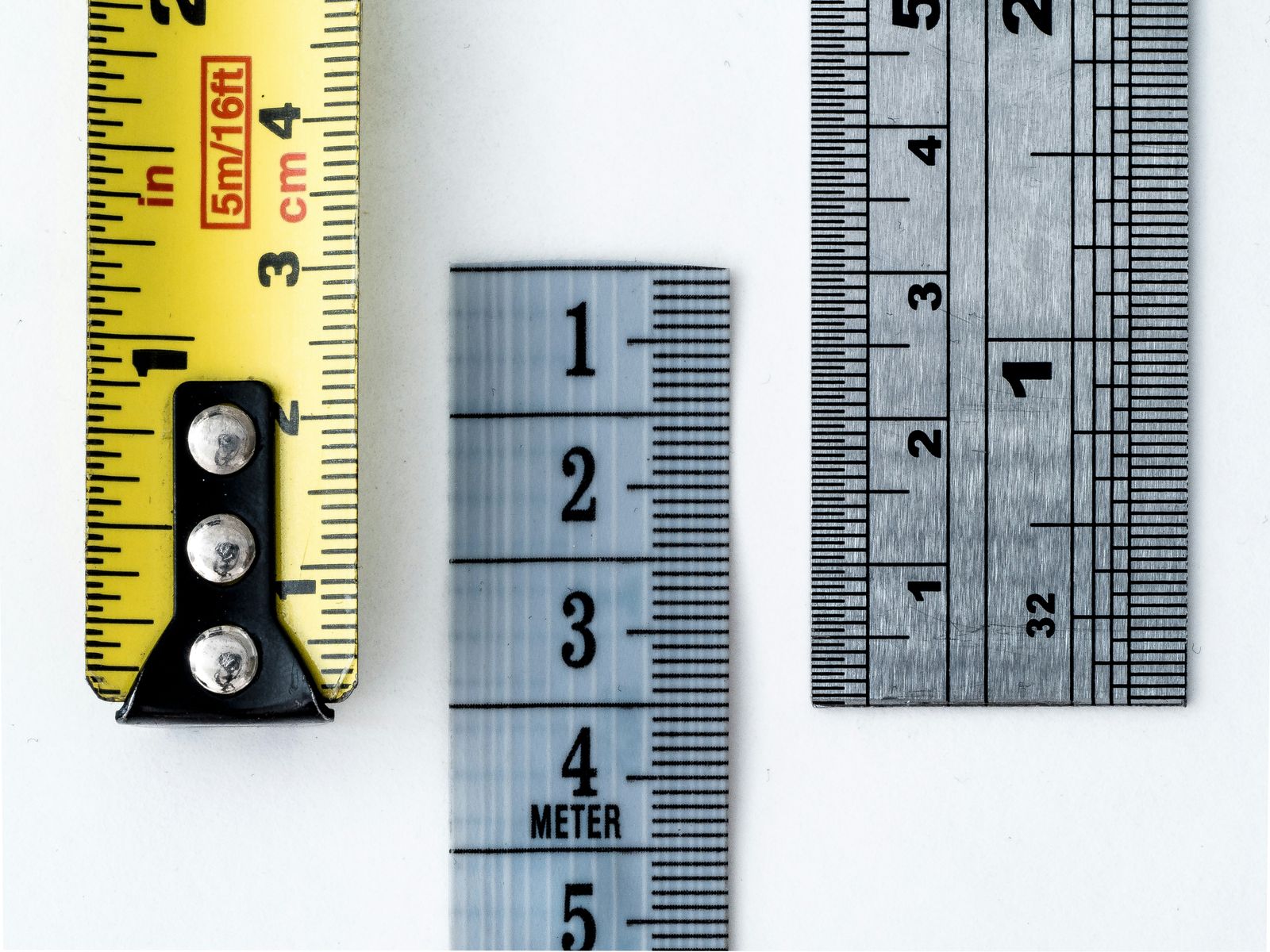 Close-up of three measuring tools: a yellow tape measure marked in inches and centimeters, a translucent ruler labeled in meters and centimeters, and a metallic ruler marked in inches with 1/32 subdivisions.