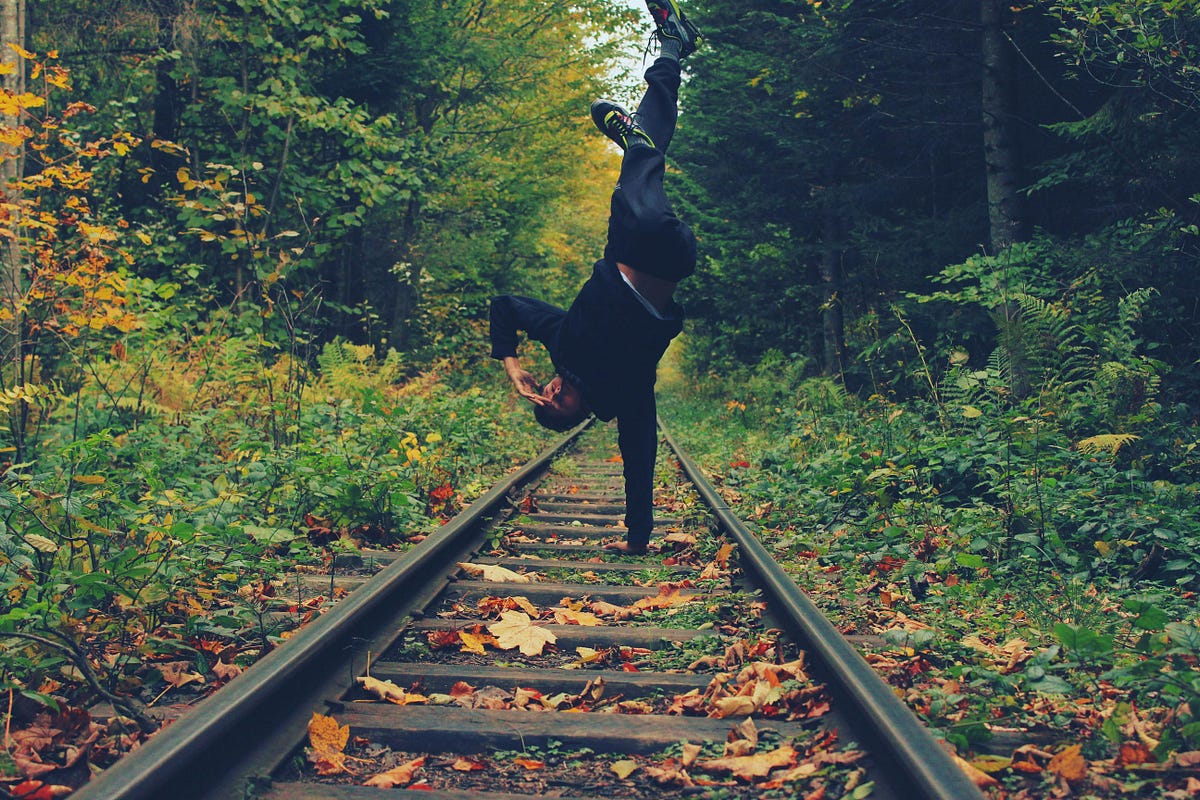 Person performing a one-handed handstand on a railroad track surrounded by dense, colorful autumn foliage