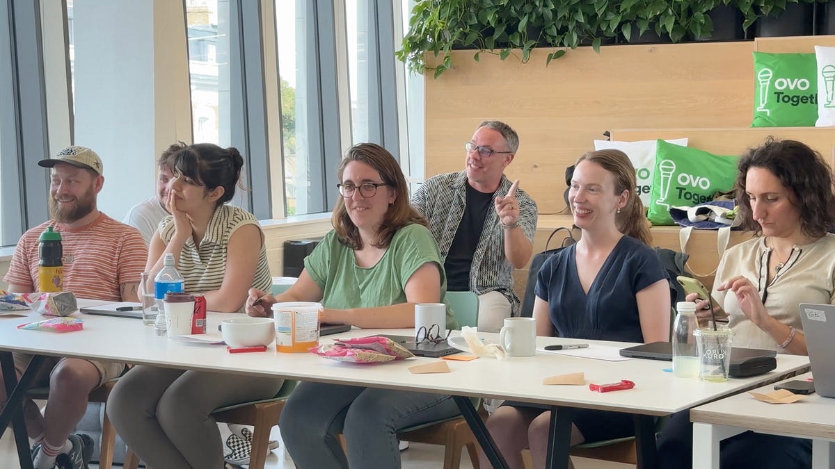 Six people sitting at a table with drinks and papers, laughing and interacting in a bright room with plants and logo cushi...