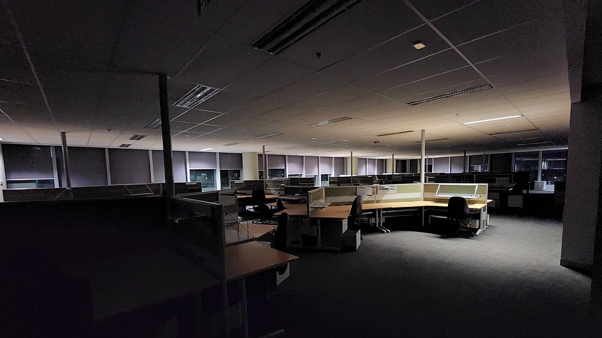 Dimly lit empty office with rows of cubicles, chairs, and computer monitors, blinds drawn on windows.