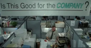 Crowded office cubicle with stacks of paper, computer, and phone. Man sits in the middle under banner reading "Is This Goo...