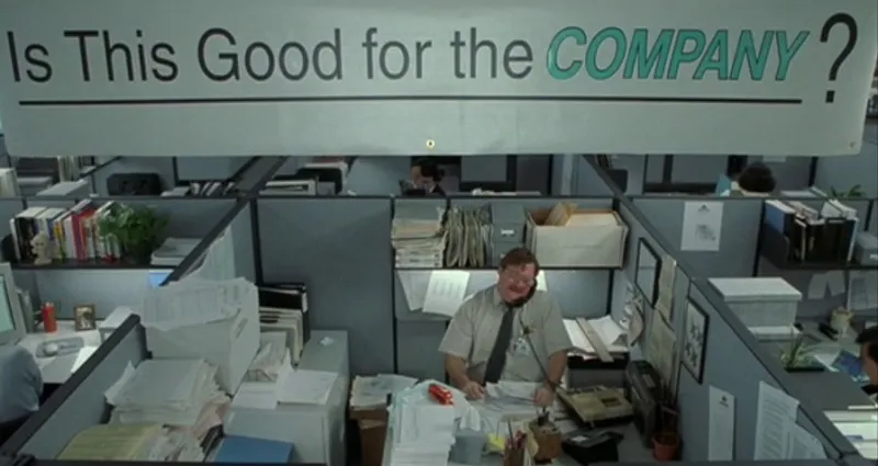 Crowded office cubicle with stacks of paper, computer, and phone. Man sits in the middle under banner reading "Is This Goo...